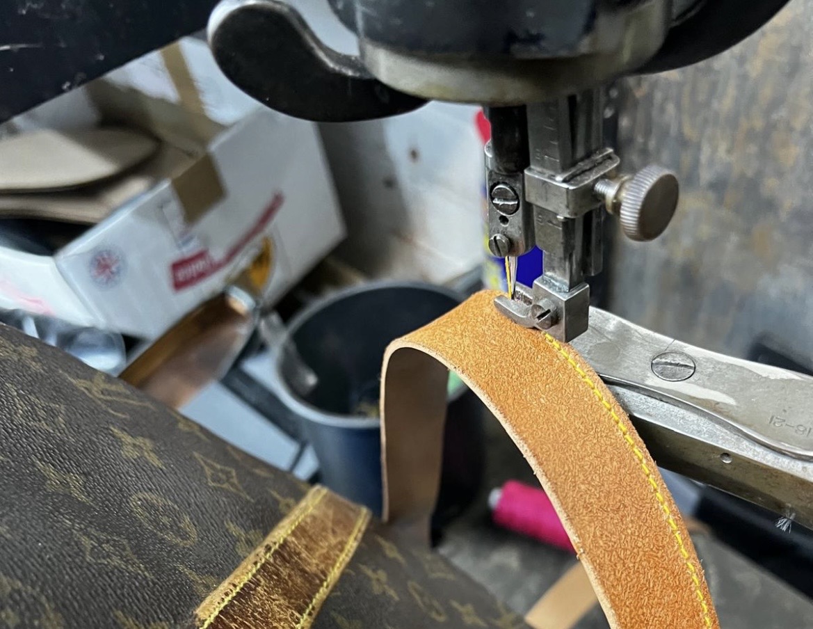 Industrial sewing machine stitching a tan leather replacement strap onto a luxury handbag.