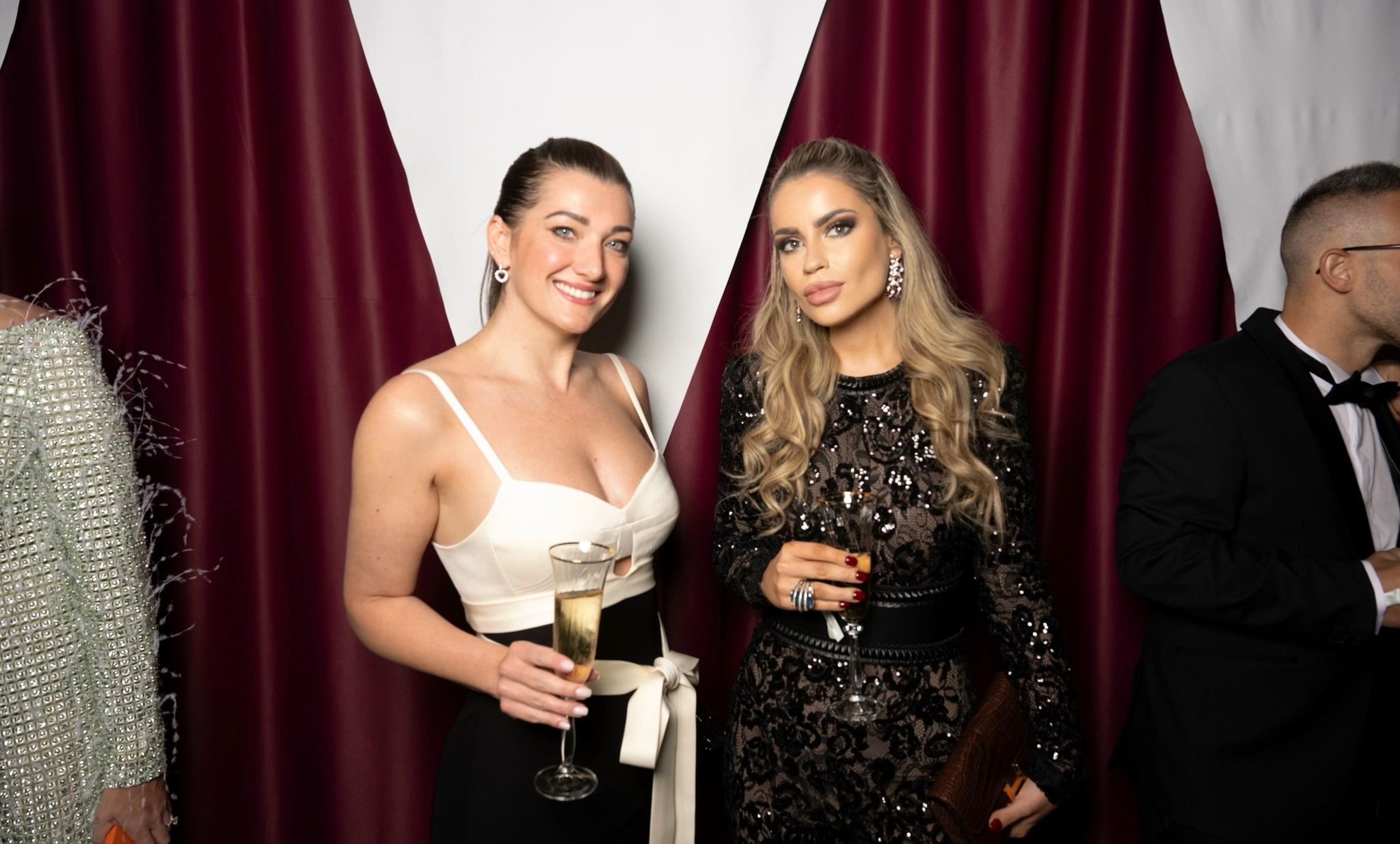 Two guests in evening wear holding champagne at a formal event with red draped backdrop