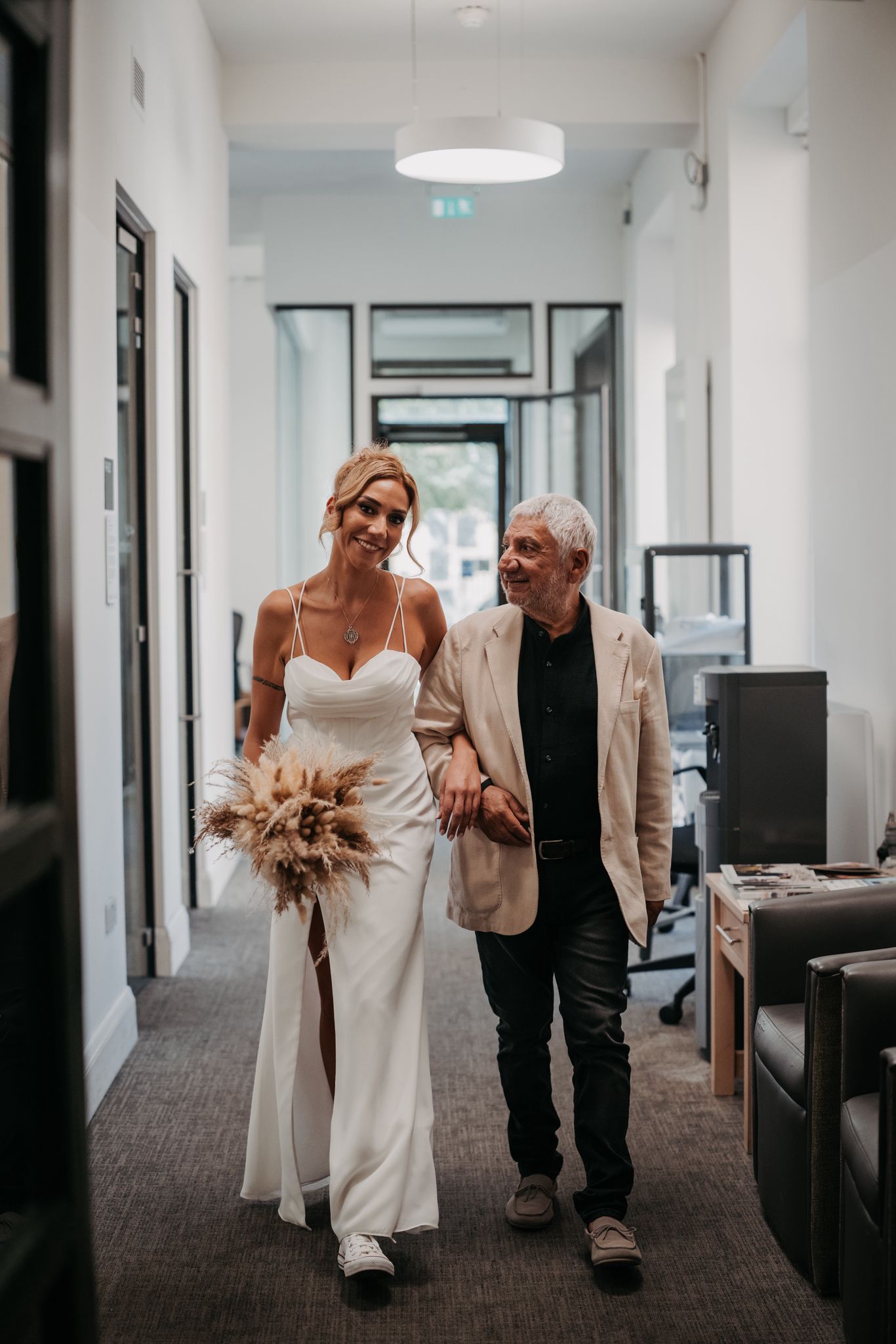 Minimalist white wedding dress with a thigh slit and sneakers, worn walking arm-in-arm indoors