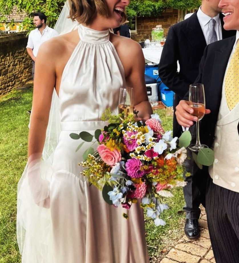 Bride in an ivory satin halter wedding dress holding a colourful bouquet at an outdoor reception