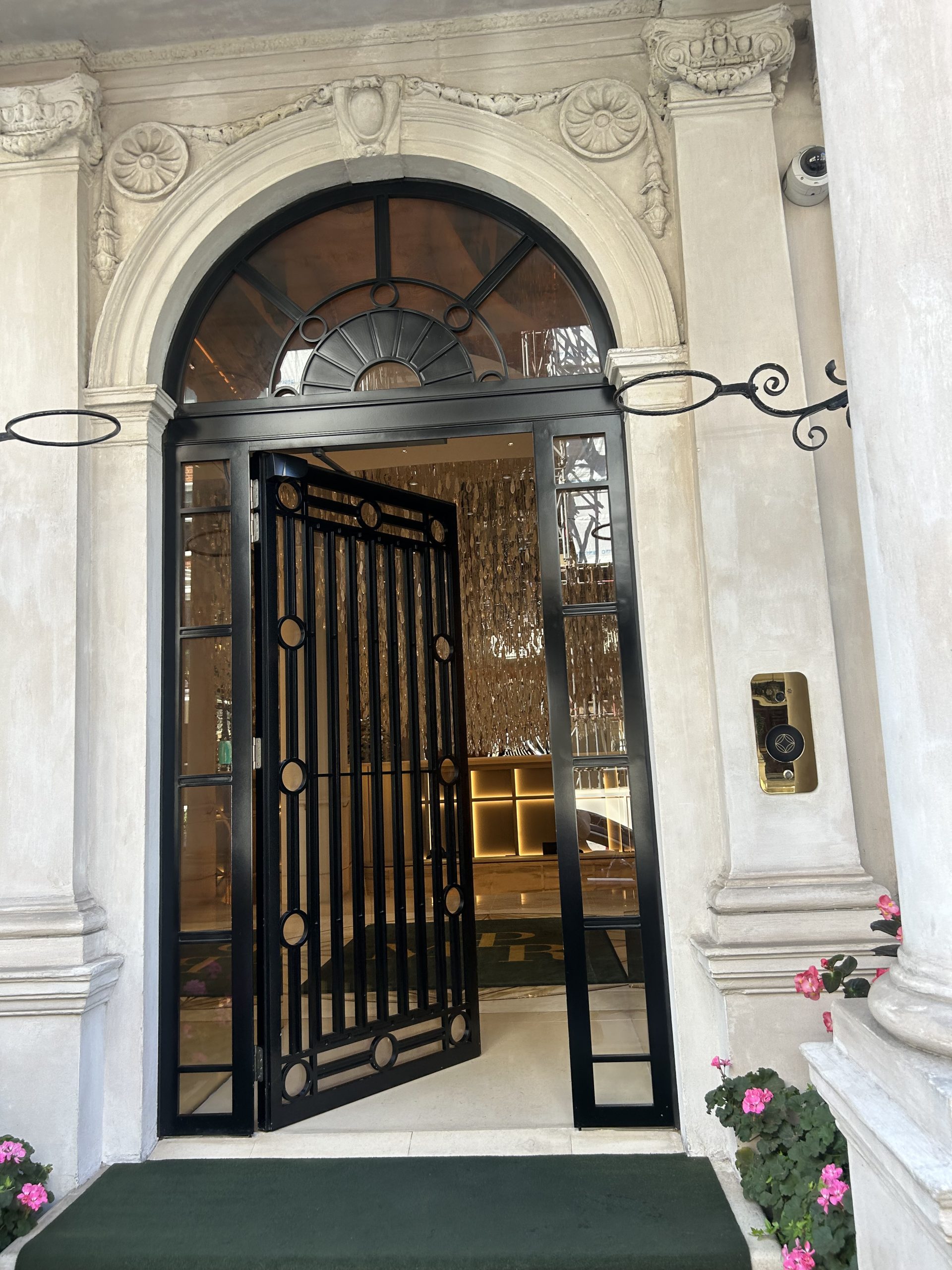 Ornate classical stone doorway with an open black metal door revealing a chandelier-lit interior.