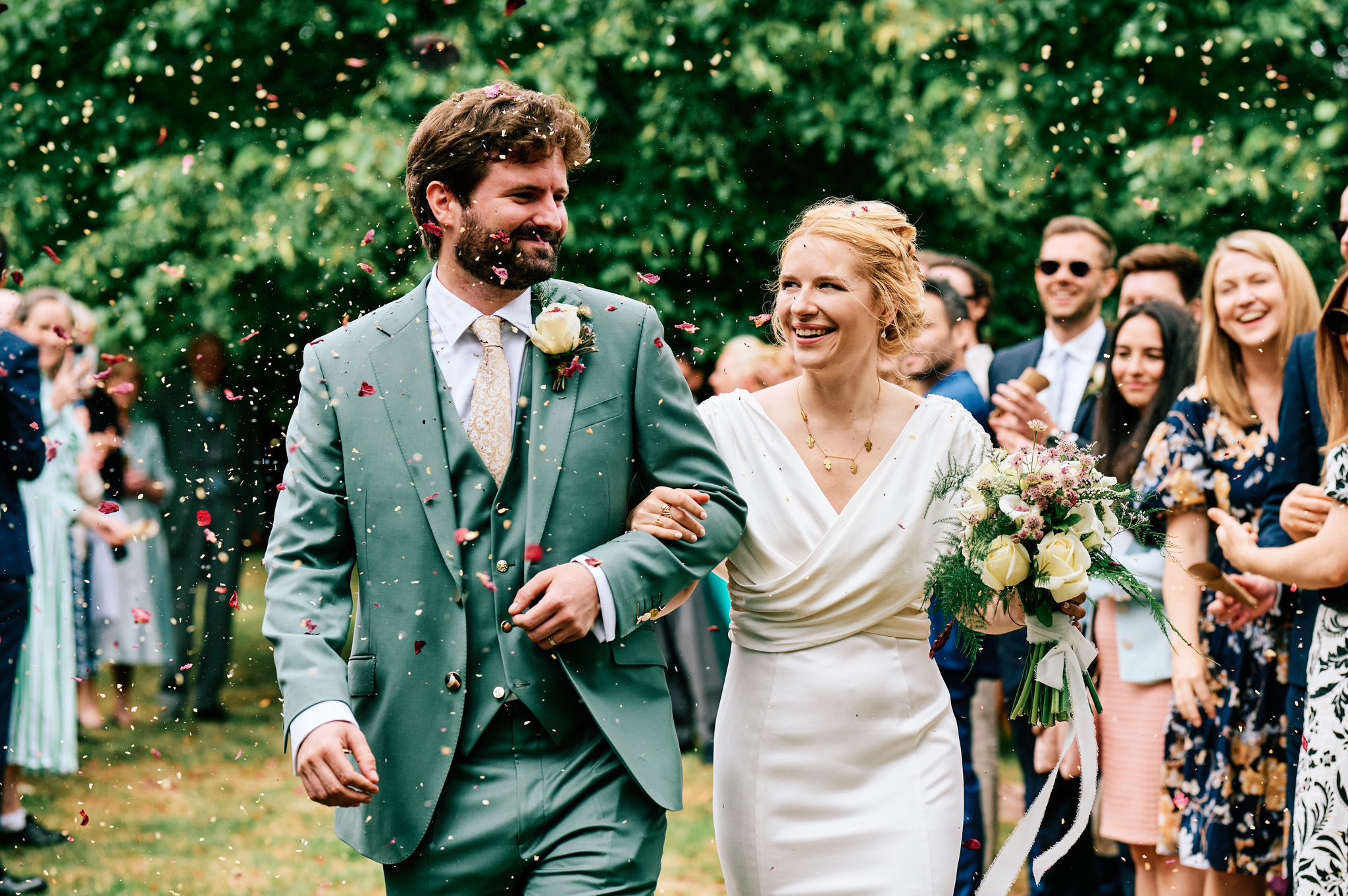 Bride in a long-sleeve wedding dress and groom in a sage green three-piece suit under confetti