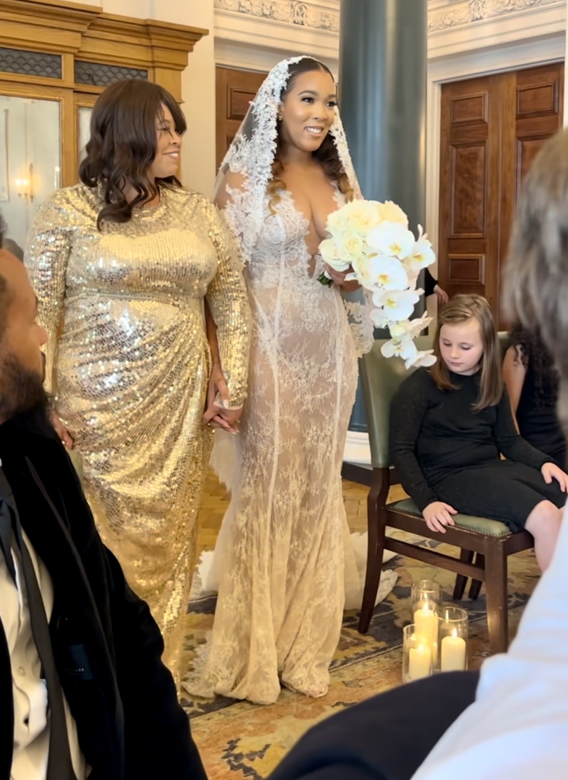 Bride in a lace mermaid wedding dress and long lace veil walking with a guest in a gold sequin gown