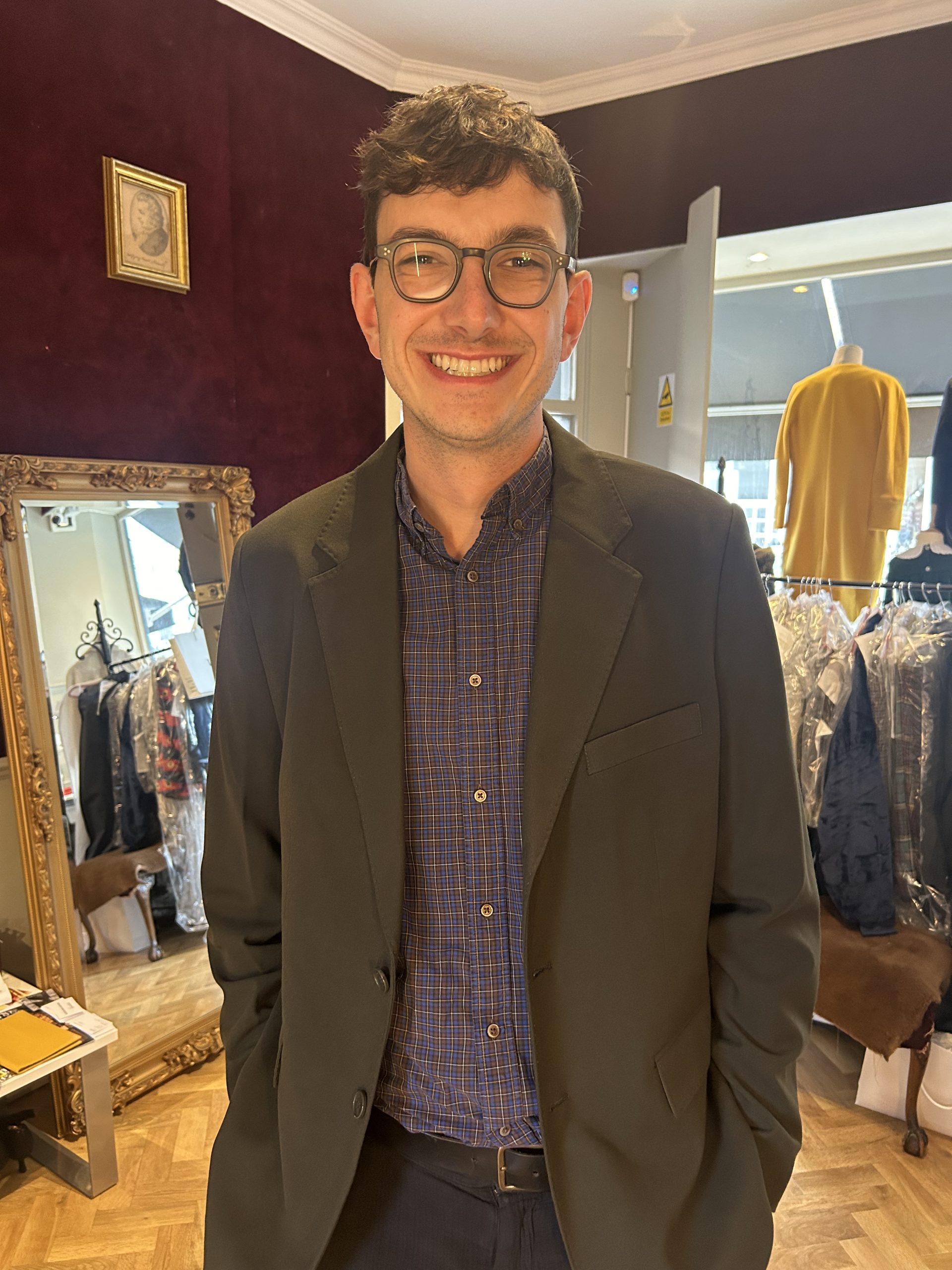 Man in a dark tailored jacket over a checked shirt, smiling in a bespoke tailoring atelier