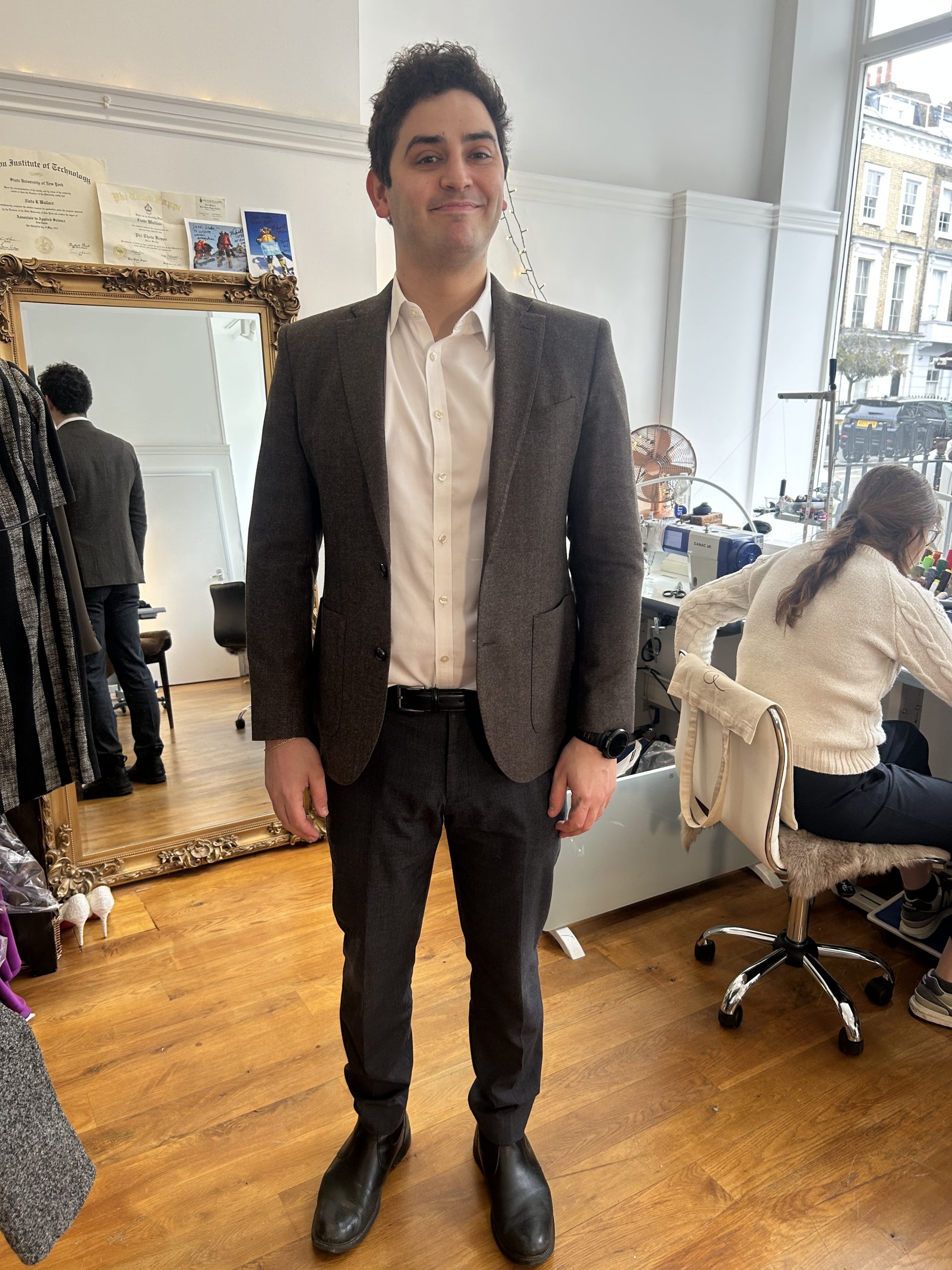 Man in a charcoal-brown textured wool jacket and tailored trousers posing in a bright bespoke atelier