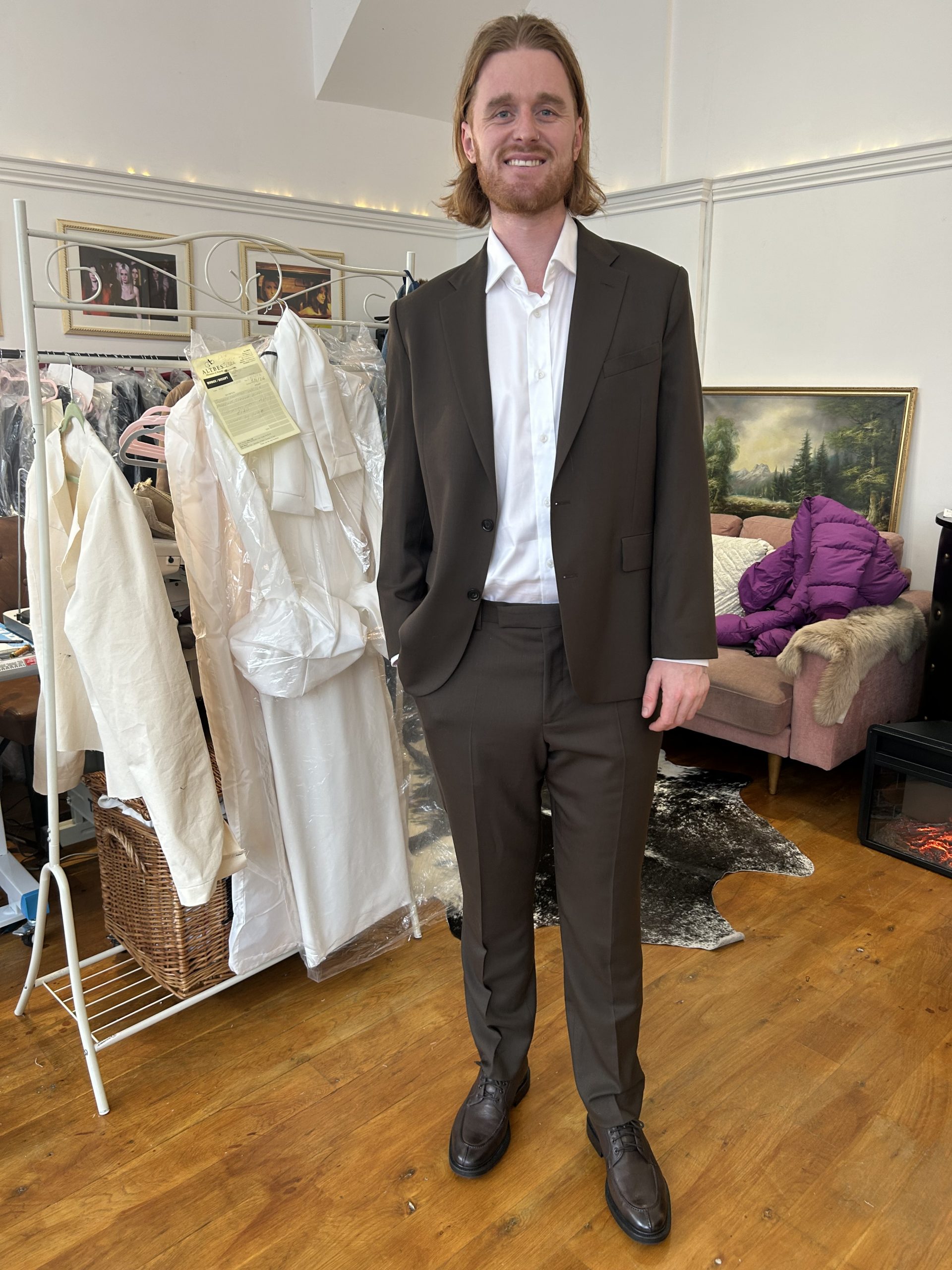 Man smiling in a chocolate-brown two-piece bespoke suit with an open-collar shirt in an atelier