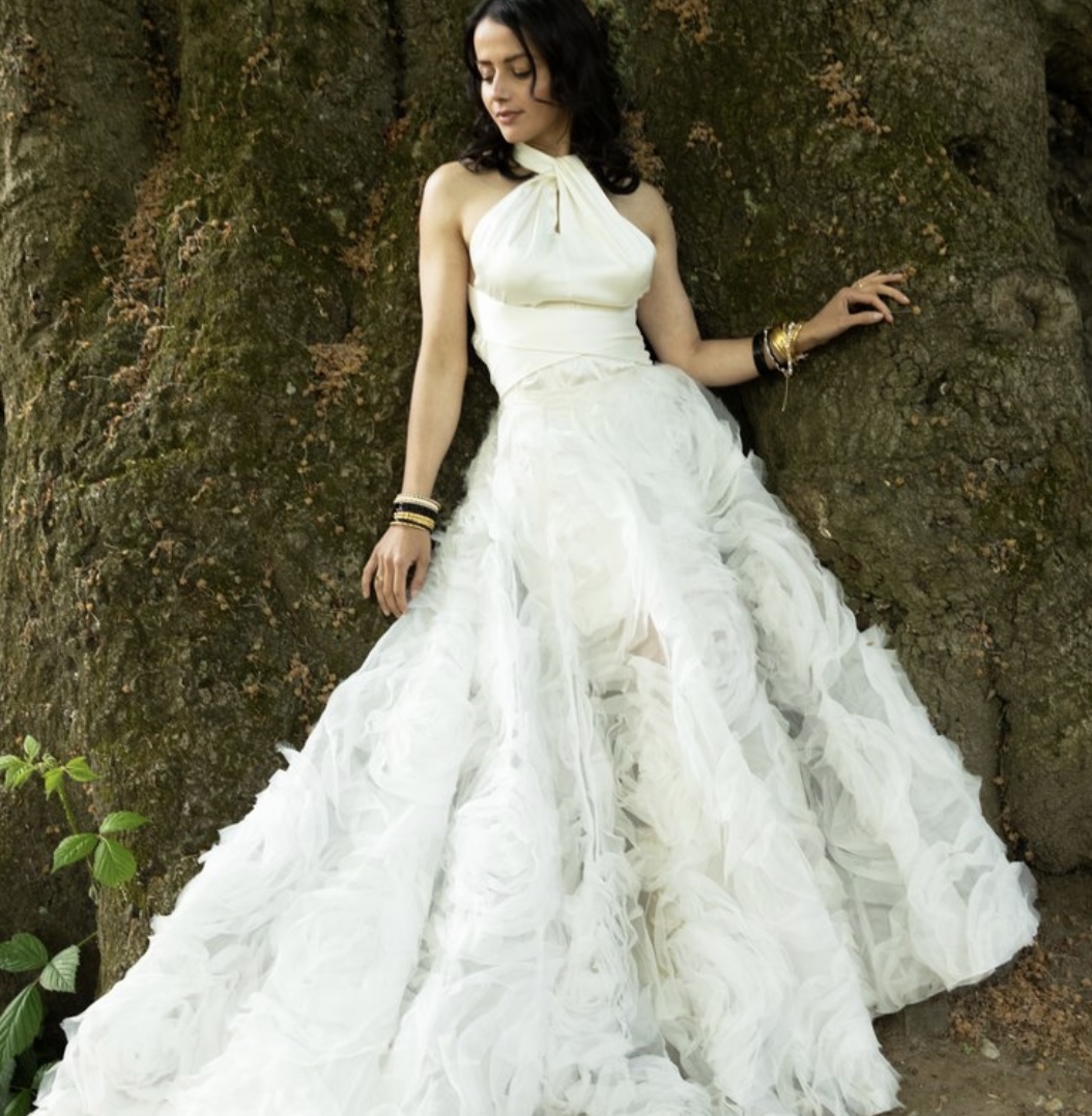 Bride in a white halter wedding gown with a ruffled tulle skirt, leaning against a mossy tree