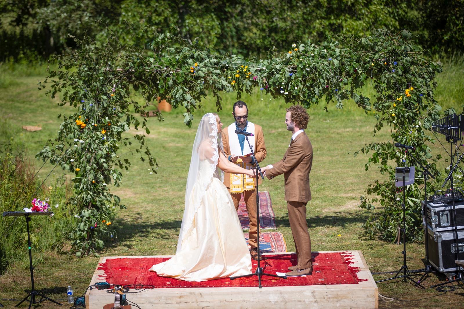 Outdoor wedding ceremony under a leafy arch; ivory A-line wedding dress and tan bespoke suit